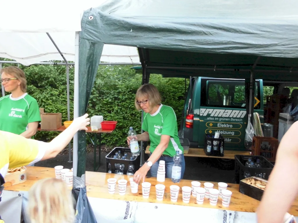 Zwei Frauen im grünen T-Shirt stehen an einem Stand auf einem Event, während sie Getränke und Snacks ausgeben. Im Hintergrund ist ein Fahrzeug mit Werbeschrift zu sehen. Der Stand ist mit Tischen und einer Markise ausgestattet, umgeben von grüner Vegetation.