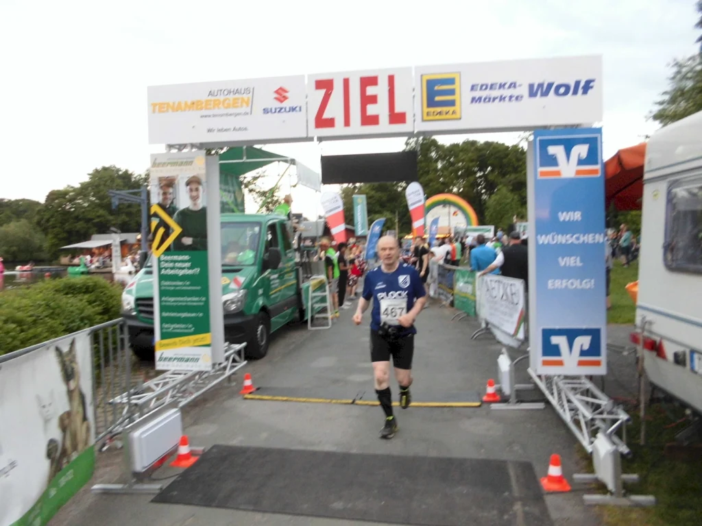 Läufer überquert die Ziellinie bei einem Wettlauf, mit Sponsorenbanner von EDEKA und anderen Firmen im Hintergrund. Die Veranstaltung findet in einer festlichen Atmosphäre statt, umgeben von Zuschauern und bunten Dekorationen.