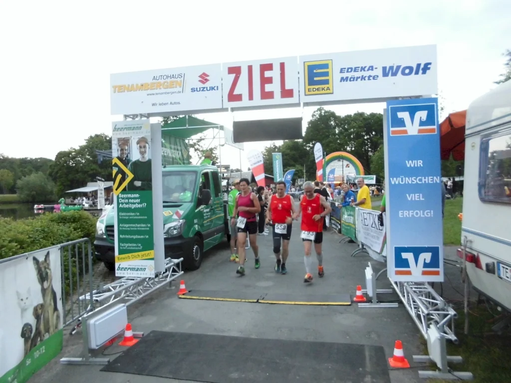 Läufer erreichen das Ziel bei einem Wettkampf, umgeben von Sponsorenbannern und Zuschauern. Im Hintergrund sind Fahrzeuge und Festzelte sichtbar.