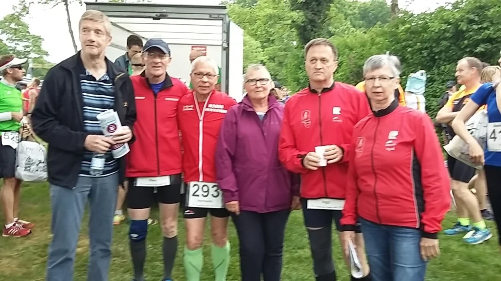 Gruppe von sechs Läufern in Sportkleidung, die sich vor dem Start eines Rennens versammeln. Sie tragen rote Jacken und zeigen Vorfreude auf die Veranstaltung. Im Hintergrund sind weitere Teilnehmer zu sehen, die sich auf das Rennen vorbereiten.