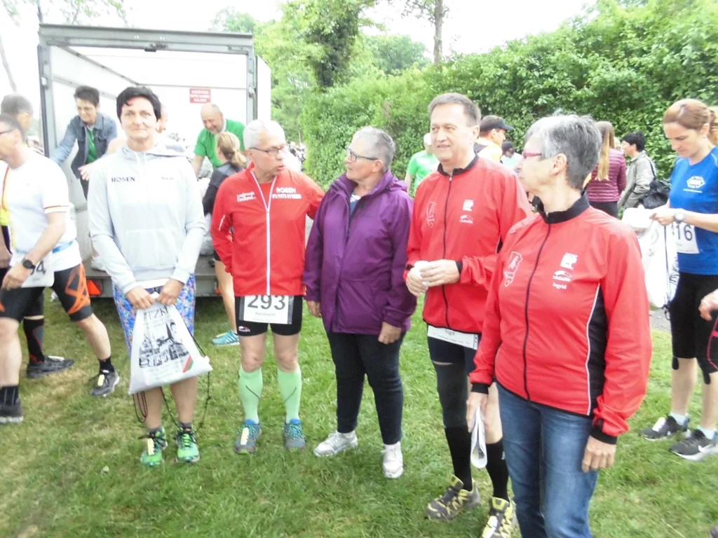 Teilnehmer eines Lauf-Events stehen in sportlicher Kleidung auf einer Wiese, während im Hintergrund weitere Personen und ein Transportfahrzeug zu sehen sind. Die Gruppe besteht aus fünf Personen, darunter Läufer in verschiedenen Outfits und mit Startnummern.