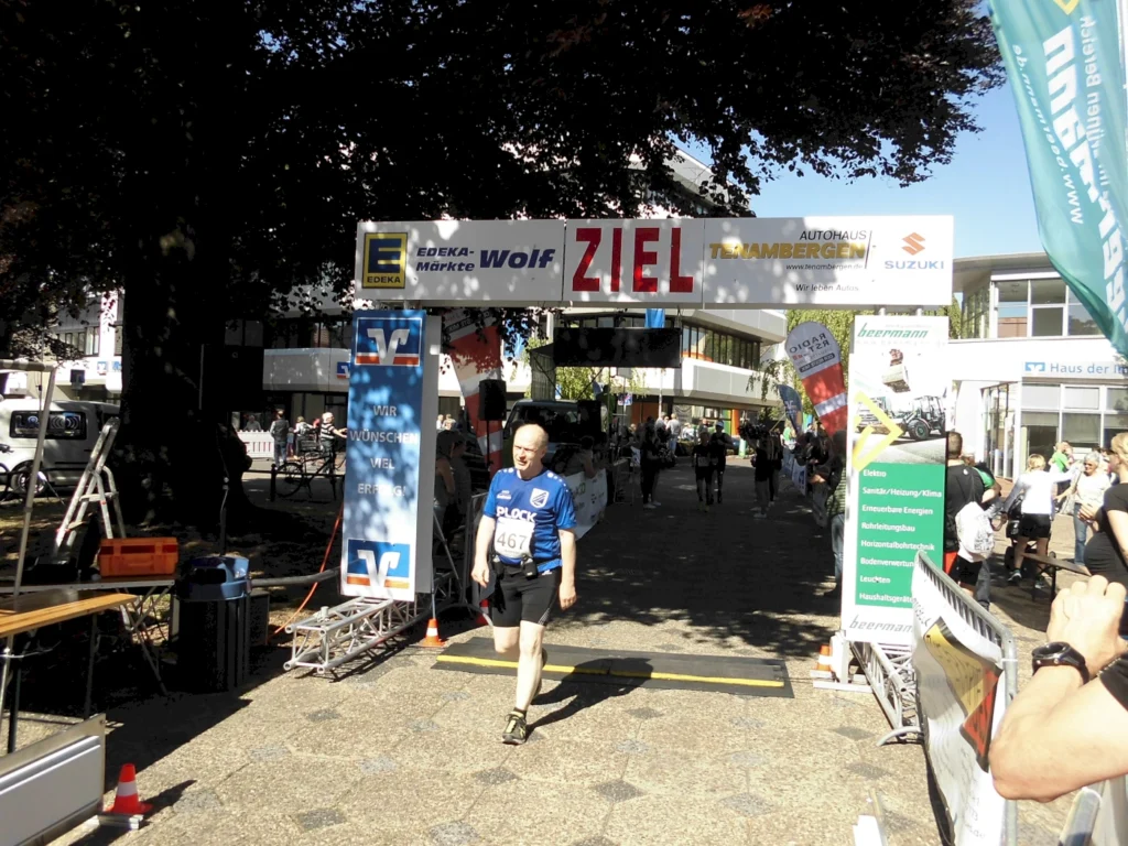 Teilnehmer eines Straßenrennens überqueren die Ziellinie unter einem großen Banner mit der Aufschrift "ZIEL". Im Hintergrund sind Zuschauer und Sponsorenbanner sichtbar, die die Veranstaltung unterstützen.