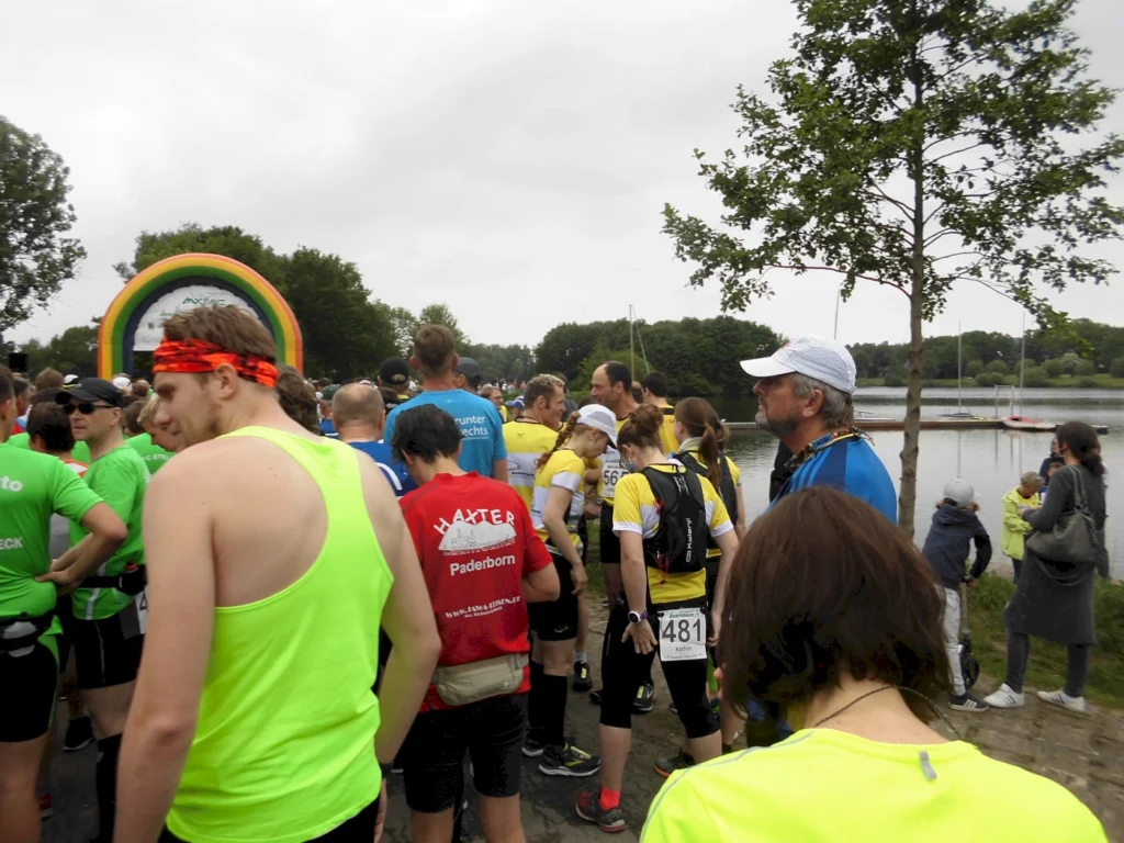 Teilnehmer eines Lauf-Events versammeln sich am Start mit einem Regenbogenbogen im Hintergrund. Die Sportler tragen bunte Laufkleidung in verschiedenen Farben, während sich eine malerische Seenlandschaft im Hintergrund erstreckt.