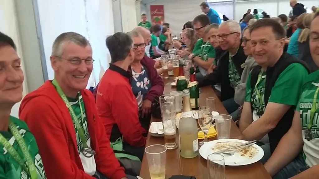 Gruppe von Menschen in einem Festzelt, die an einem Tisch sitzen, mit Getränken und Essen vor sich. Die Teilnehmer tragen grüne Shirts und Medaillen, was auf eine sportliche Veranstaltung hindeutet. Die Atmosphäre ist gesellig und feierlich.