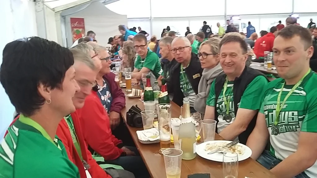 Gruppengespräch bei einer Veranstaltung in einem Zelt, mit Teilnehmern in grünen T-Shirts, Essen und Getränken auf dem Tisch. Die Atmosphäre ist gesellig und lebhaft.