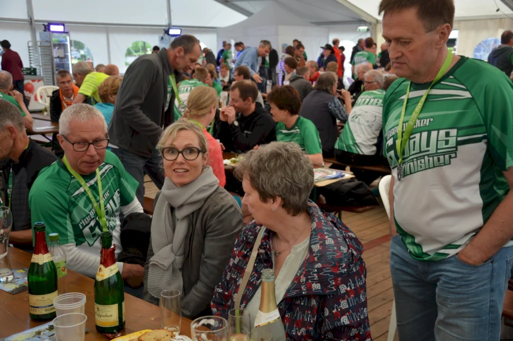 Gruppe von Menschen in einem Festzelt, die an Tischen sitzen und sich unterhalten. Einige tragen grüne Sportshirts, während Getränke wie Sekt und Bier auf den Tischen stehen. Die Atmosphäre wirkt gesellig und festlich, ideal für ein gemeinsames Event oder eine Feier.