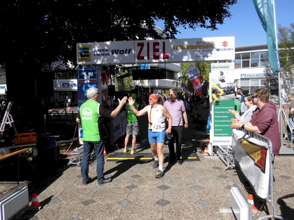 Läufer überquert die Ziellinie eines Wettkampfs, umgeben von Zuschauern und Unterstützern. Im Hintergrund sind Sponsorenbanner und eine festliche Atmosphäre sichtbar.