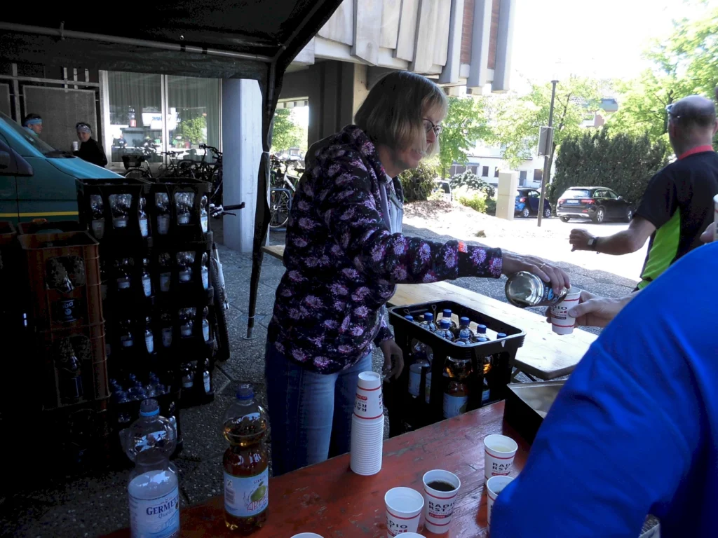 Eine Frau füllt Getränke in einen Becher an einem Stand, umgeben von Wasserflaschen und anderen Getränken. Im Hintergrund sind Fahrräder und Menschen zu sehen, die an einer Veranstaltung teilnehmen.