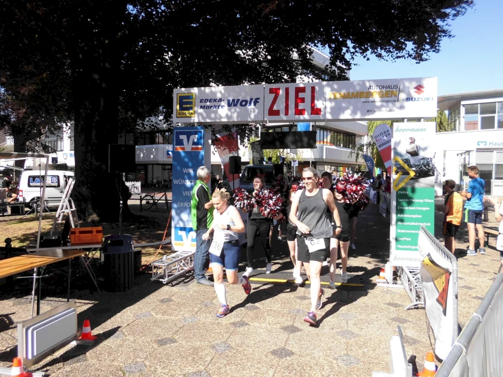 Läufer überqueren die Ziellinie bei einem Sportevent, umgeben von jubelnden Cheerleadern und Sponsorenbanner. Sonniger Tag mit festlicher Atmosphäre.