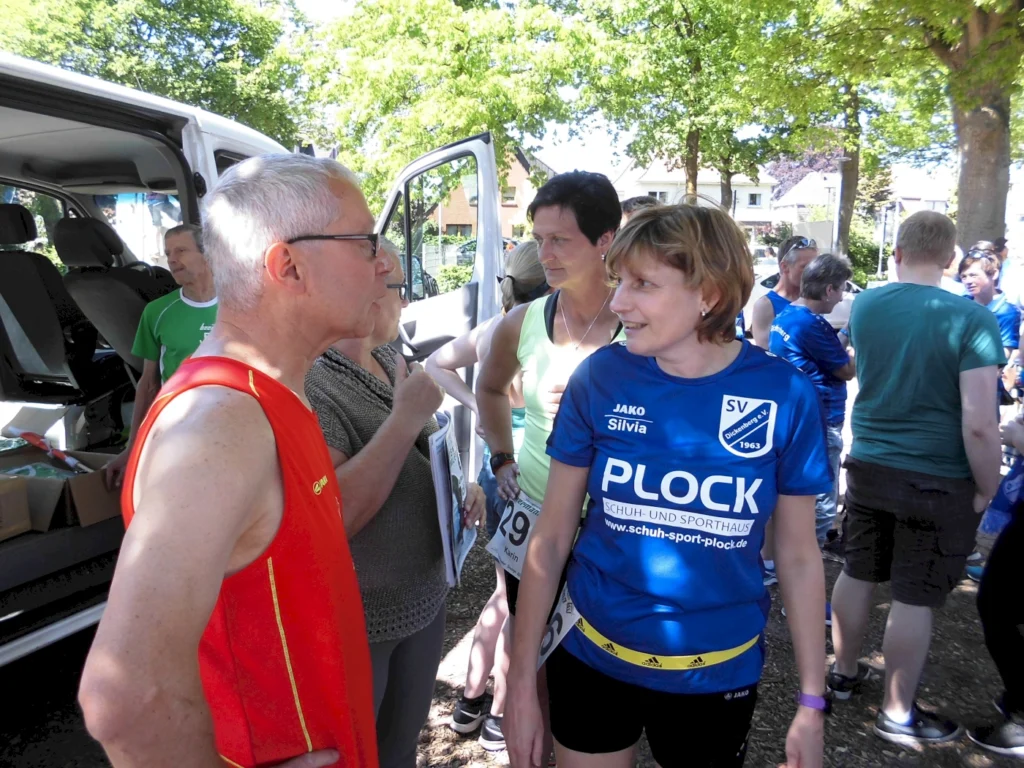 Teilnehmer eines Lauf-Events im Freien, die sich vor dem Start unterhalten. Im Vordergrund stehen zwei Personen, eine Frau in einem blauen Sporttrikot mit dem Logo des SV Dielingen und ein Mann in einem roten Tanktop. Im Hintergrund sind weitere Teilnehmer und ein Transporter zu sehen, umgeben von Bäumen und sonnigem Wetter.