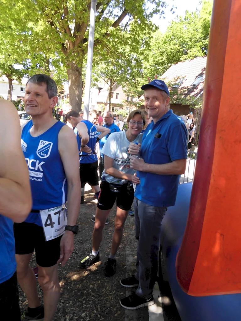 Gruppenszene bei einem Laufereignis, Teilnehmer in blauen Trikots stehen aufgereiht, darunter eine Frau mit Wasserflasche. Im Hintergrund sind Bäume und Zuschauer zu sehen, die Atmosphäre ist festlich und sportlich.