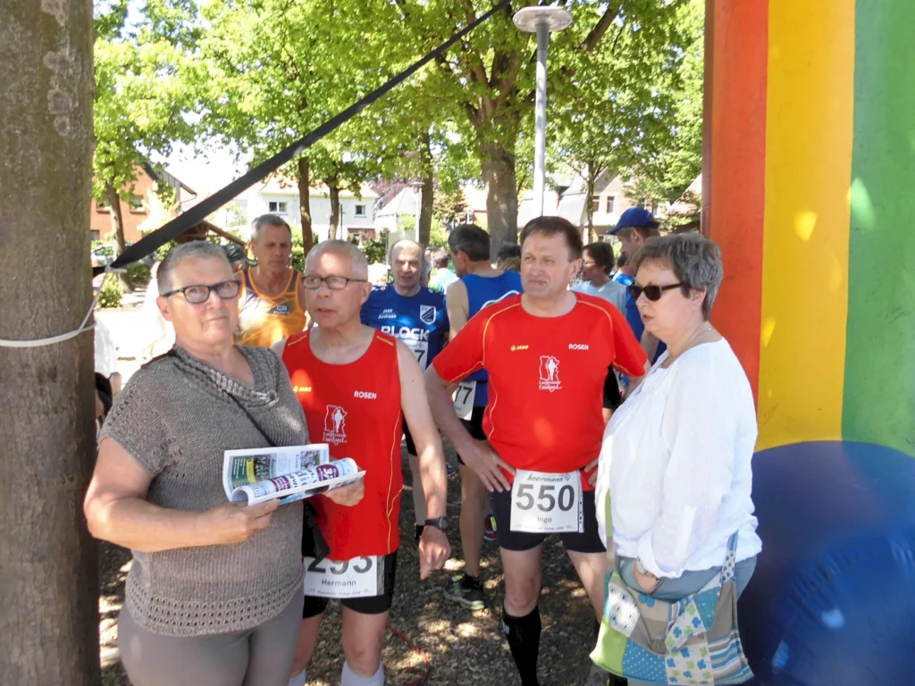 Teilnehmer des Laufwettbewerbs stehen unter Bäumen, während sie sich auf den Start vorbereiten. Im Vordergrund sind eine Frau mit Brille und ein Mann in einem roten Trikot zu sehen, die einen Flyer halten. Im Hintergrund sind weitere Läufer in bunten Trikots sichtbar. Die Szenerie ist hell und sonnig, umgeben von Grün.