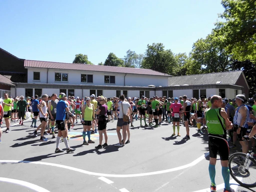 Gruppierung von Sportlern in bunten Trikots auf einem Platz vor einem Gebäude, während eines Lauf-Events bei sonnigem Wetter.