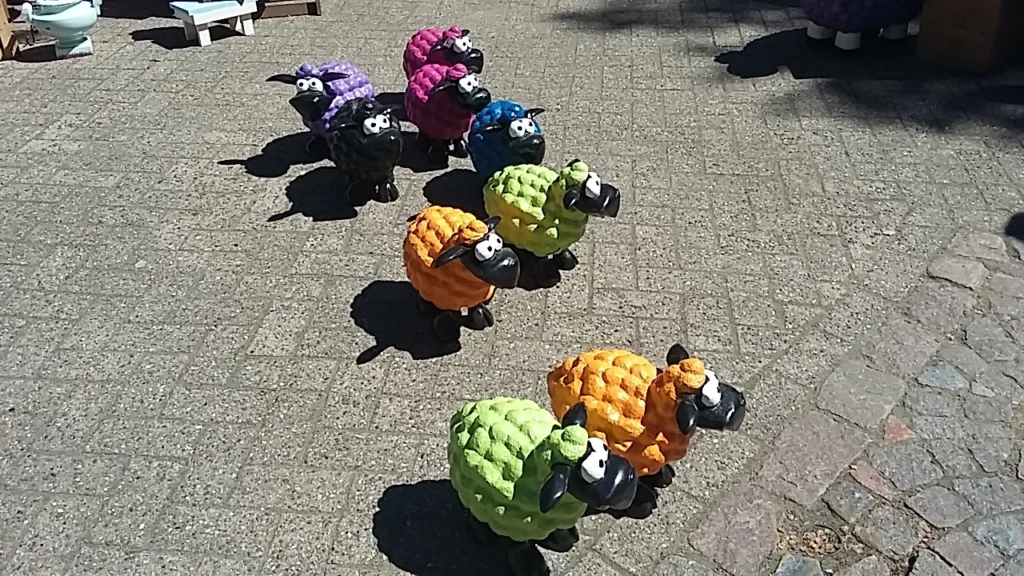 Bunte, dekorative Schafe aus Kunststoff in verschiedenen Farben (lila, schwarz, blau, grün, orange) auf einem Gehweg. Ideal für Garten- und Landschaftsgestaltung.