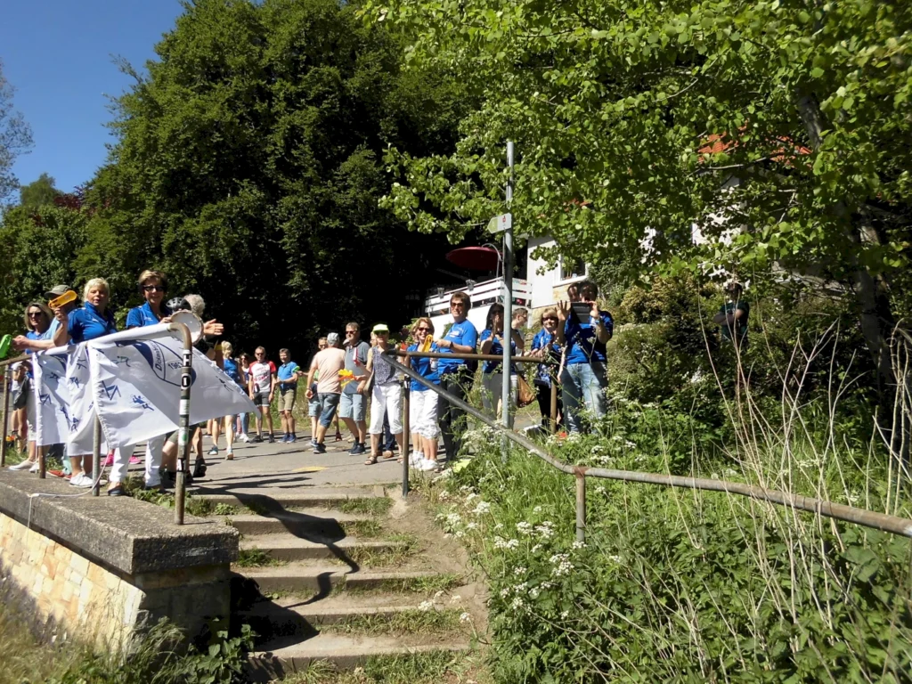 Menschenmenge auf einem Wanderweg, die in blauen T-Shirts feiert, umgeben von grüner Natur. Im Vordergrund stehen Teilnehmer mit einer Fahne, während im Hintergrund weitere Wanderer zu sehen sind. Ideal für Veranstaltungen im Freien oder Wandergruppen.