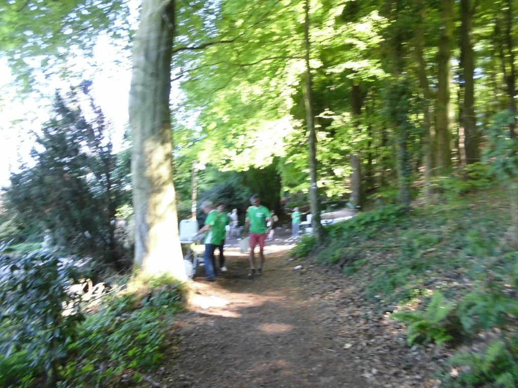 Zwei Personen in grünen T-Shirts gehen auf einem Waldweg, umgeben von üppigem Laub und Bäumen. Im Hintergrund sind weitere Menschen und ein Fahrzeug zu sehen.