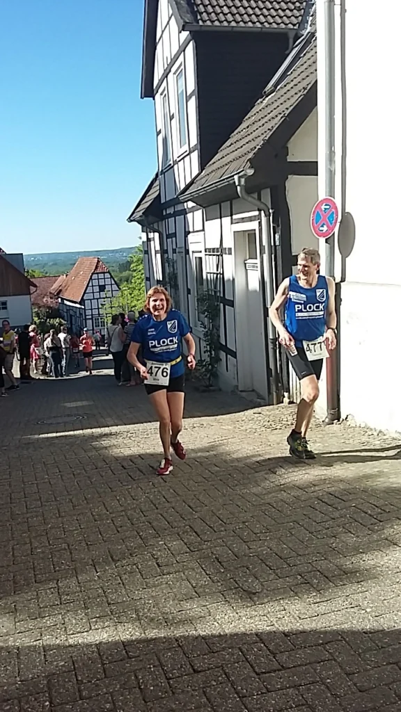 Läufer bei einem Wettkampf in einer malerischen Stadt, mit historischen Fachwerkhäusern im Hintergrund. Die Teilnehmer tragen Trikots mit der Aufschrift "PLOCK" und Startnummern. Zuschauer stehen entlang der Strecke und unterstützen die Sportler.