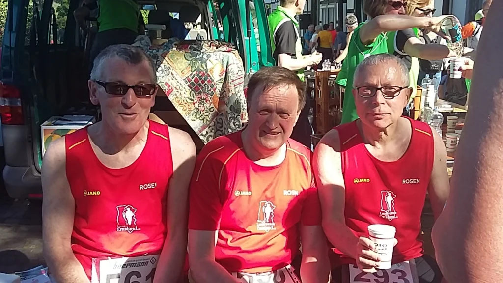 Drei männliche Läufer in roten Trikots sitzen entspannt nach einem Wettkampf. Sie halten Becher in der Hand und sind von einem festlichen Hintergrund umgeben.
