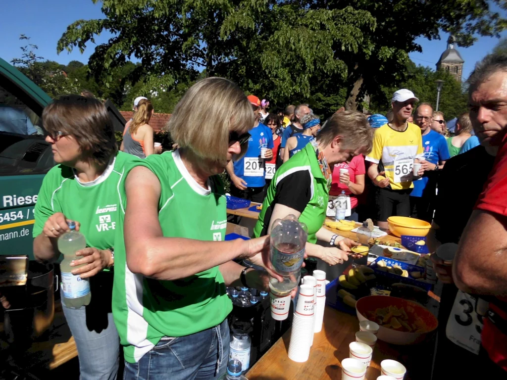 Zwei Helferinnen in grünen Trikots servieren Wasser und Snacks an einem Verpflegungstisch während eines Laufevents, umgeben von Teilnehmern in Sportkleidung.