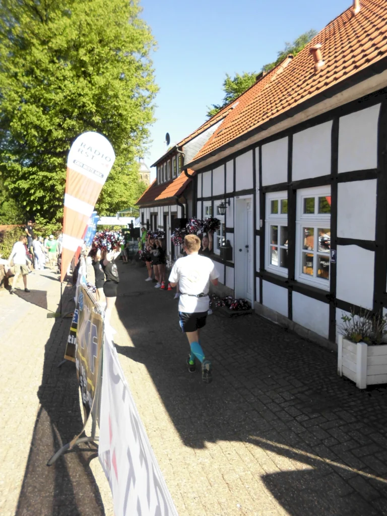 Läufer in Sportbekleidung auf einer Straße mit Fachwerkhäusern, umgeben von grünen Bäumen und einer fröhlichen Atmosphäre, während einer Veranstaltung oder einem Wettkampf.