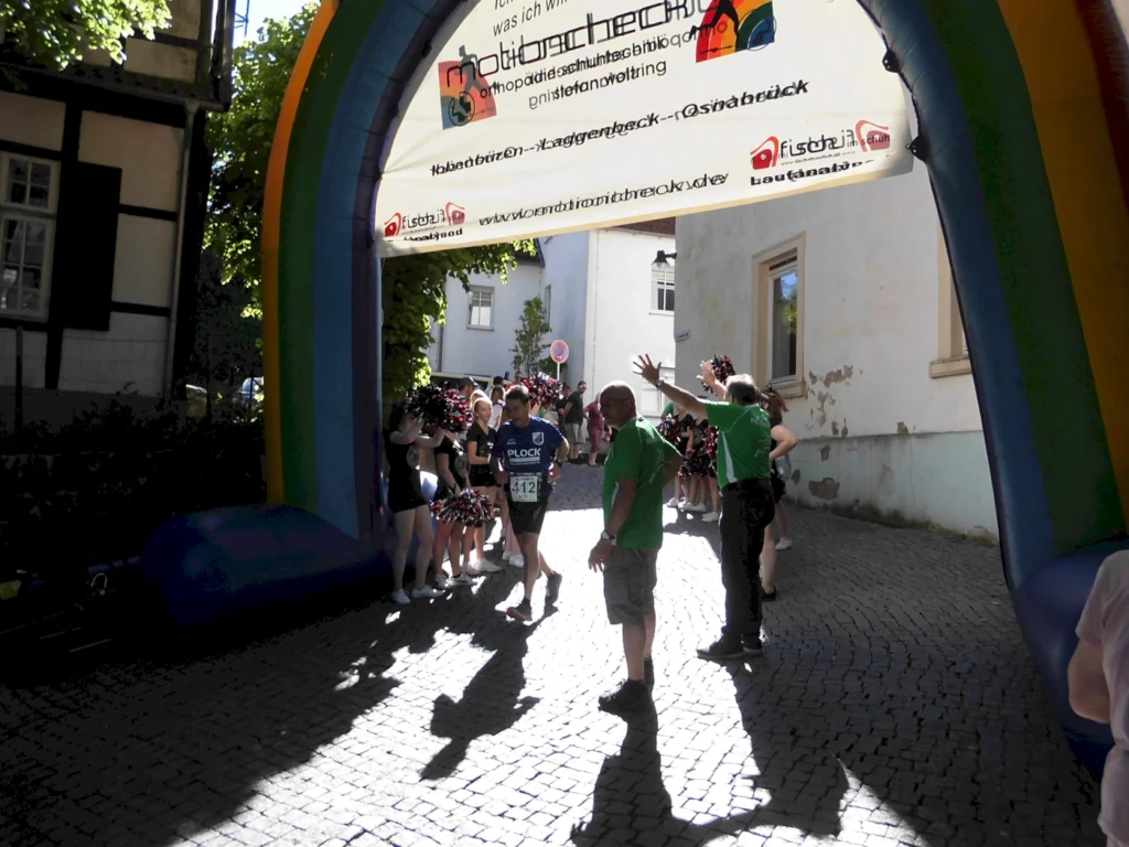 Läufer überquert das Ziel unter einem bunten Bogen bei einer Sportveranstaltung, umgeben von jubelnden Zuschauern und Helfern in grünen Shirts.