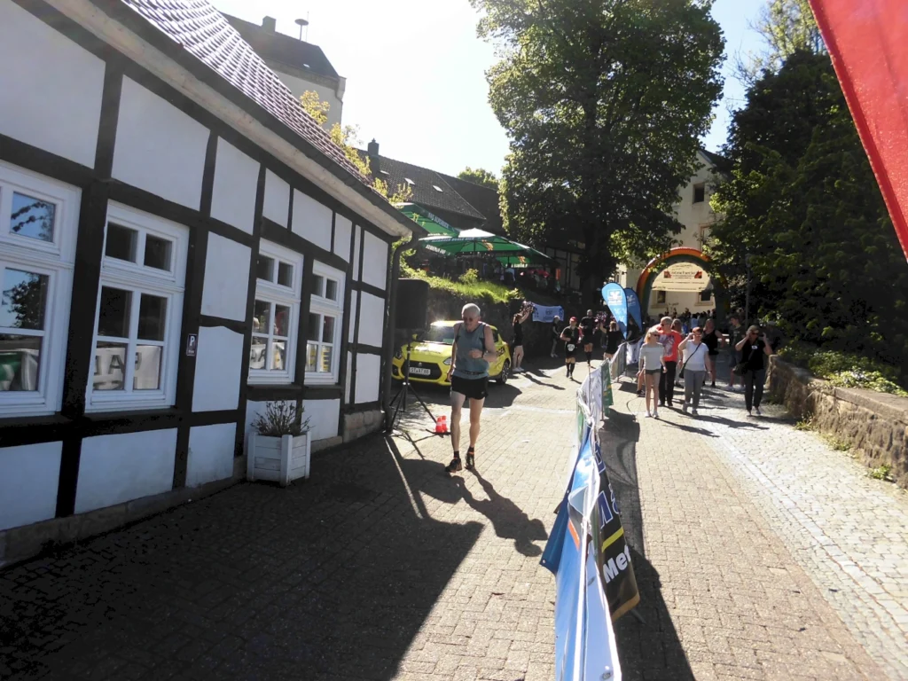 Läufer bei einem Wettkampf in einer malerischen Straße mit Fachwerkhäusern, umgeben von Zuschauern und bunten Bannern. Sonniger Tag, lebhafte Atmosphäre und lokale Gastronomie im Hintergrund.