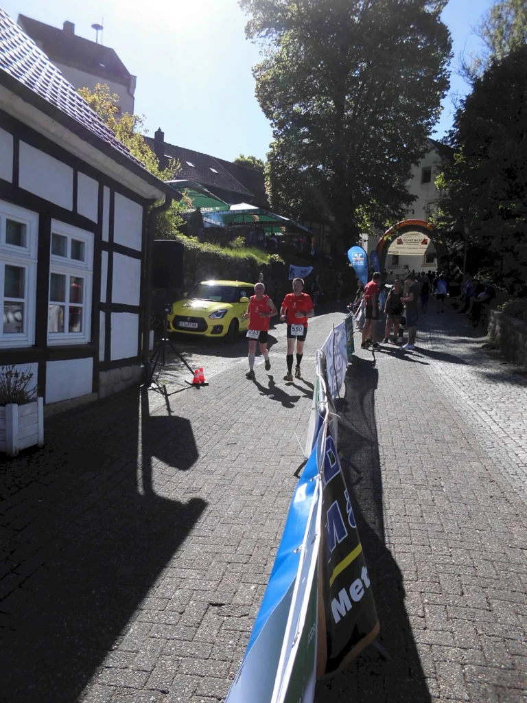 Zwei Läufer in roten T-Shirts nehmen an einem Straßenlauf teil, während Zuschauer an der Strecke stehen. Im Hintergrund ist ein gelbes Auto und eine festliche Atmosphäre mit bunten Bannern und Bäumen zu sehen.