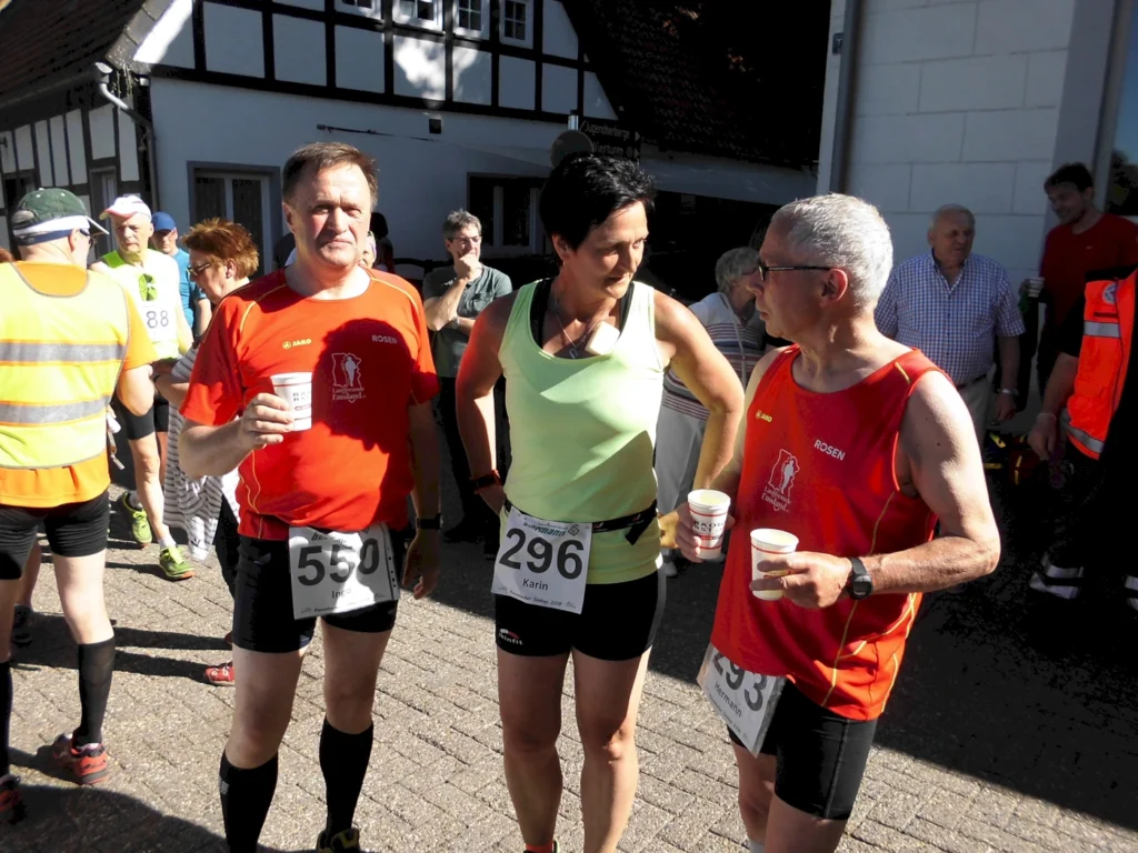 Drei Teilnehmer eines Laufwettbewerbs stehen in sportlicher Kleidung zusammen, während sie Getränke halten. Im Hintergrund sind weitere Läufer und Zuschauer zu sehen, die an einer Veranstaltung im Freien teilnehmen.