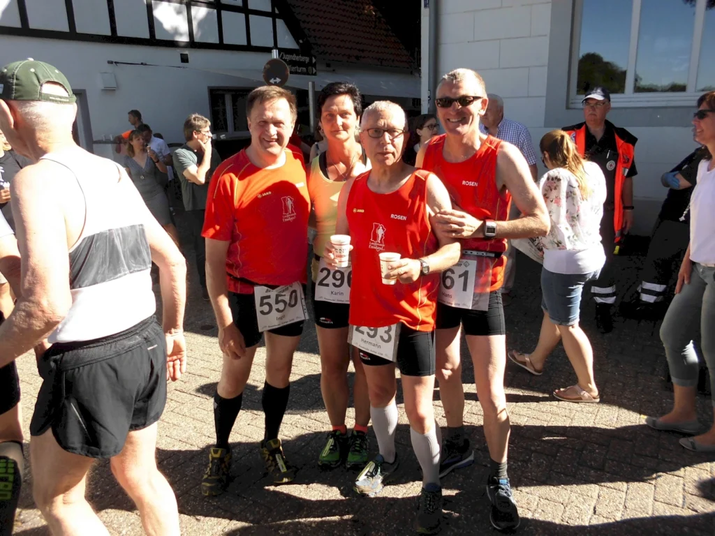 Vier Läufer in roten Trikots posieren lächelnd nach einem Wettkampf, während Zuschauer und Veranstalter im Hintergrund zu sehen sind. Die Läufer tragen Startnummern und halten Getränke in der Hand.