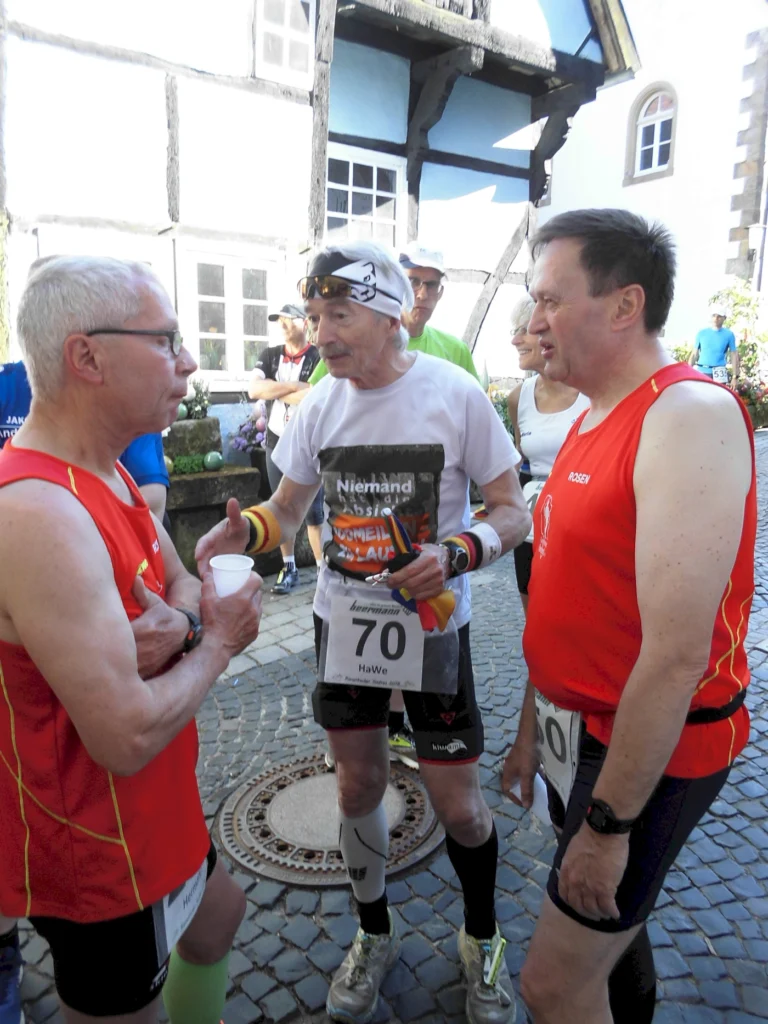 Drei Männer im Sportoutfit stehen im Gespräch, während sie sich auf einen Lauf vorbereiten. Der ältere Mann in der Mitte trägt ein T-Shirt mit der Aufschrift „Niemand hält mich auf“ und hält eine kleine Tasse in der Hand. Die anderen beiden Männer, einer in rotem Shirt und der andere in einem weißen und roten Outfit, scheinen interessiert zuzuhören. Im Hintergrund sind andere Teilnehmer und ein historisches Gebäude zu sehen.