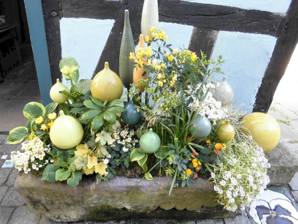 Bunte Keramikkugeln und blühende Pflanzen in einem bepflanzten Behälter vor einem Fachwerkhaus. Die Kombination aus verschiedenen Grüntönen und gelben Blumen schafft eine ansprechende Gartenlandschaft.