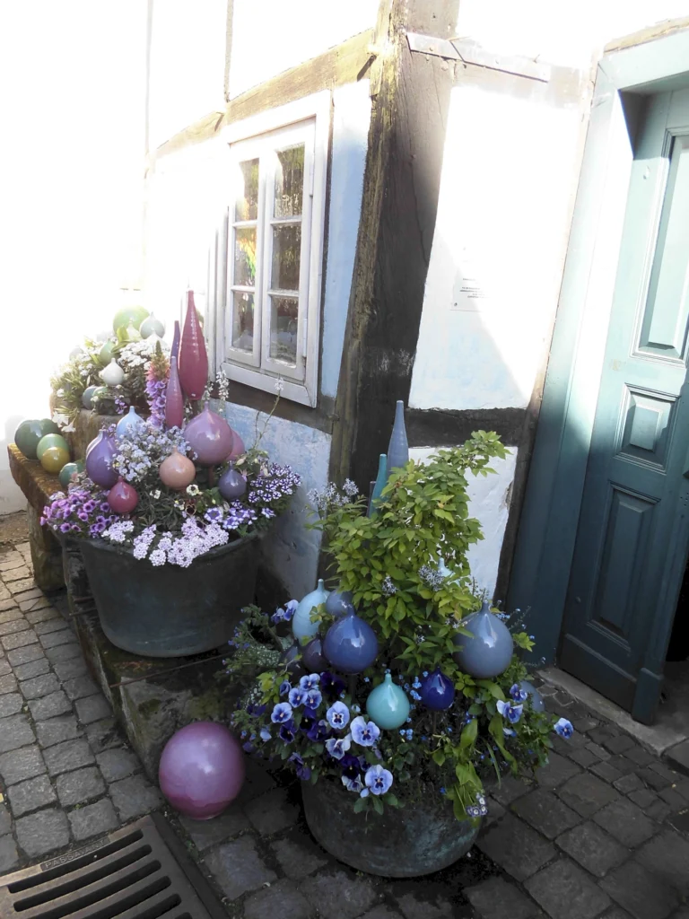 Bunte Blumentöpfe mit violetten und blauen Blumen, dekoriert mit farbigen Kugeln, neben einem historischen Fachwerkhaus und einer blauen Tür. Ideal für Gartendekoration und kreative Außenansichten.