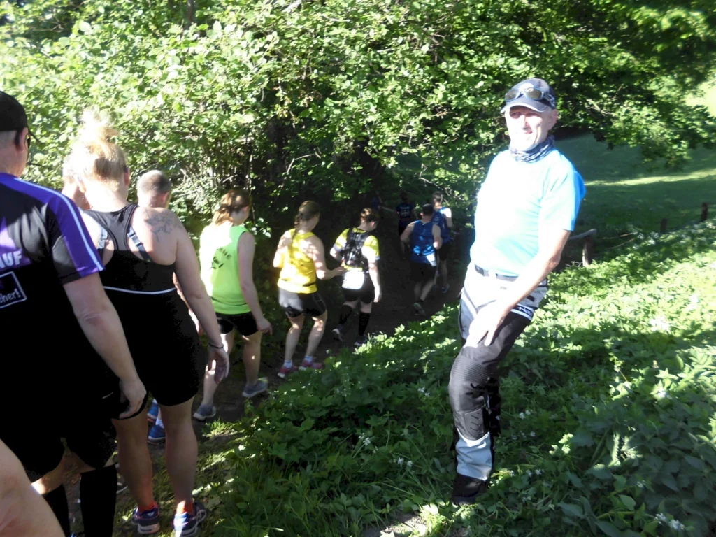 Eine Gruppe von Wanderern in Sportbekleidung, die einem schmalen Pfad durch grünes Gestrüpp folgt. Im Vordergrund steht ein Mann in blauer Sportkleidung und Sonnenbrille, während die anderen Teilnehmer in bunten T-Shirts hinter ihm gehen. Die Szene zeigt eine aktive Outdoor-Aktivität in der Natur.