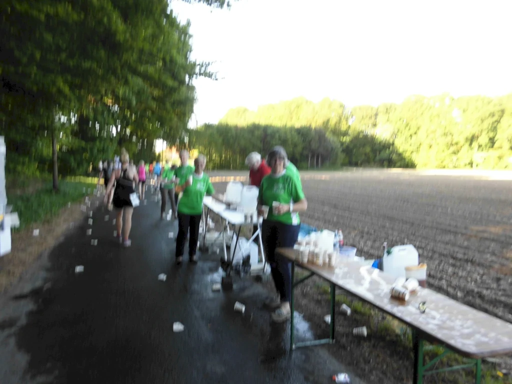 Teilnehmer eines Lauf-Events gehen an einer Verpflegungsstation vorbei, an der Freiwillige in grünen T-Shirts Getränke und Snacks anbieten. Im Hintergrund sind Bäume und ein Acker zu sehen.