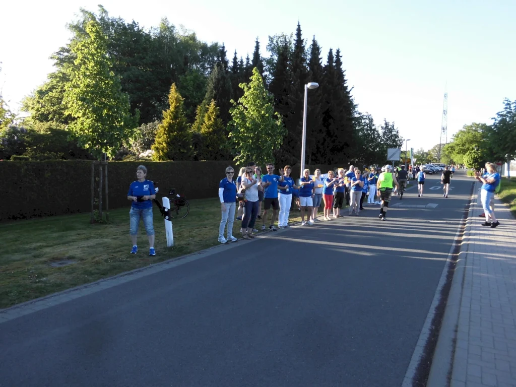 Menschenmenge in blauen T-Shirts applaudiert einem Läufer auf einer Straße, umgeben von Bäumen und einem klaren Himmel, während einer Laufveranstaltung.