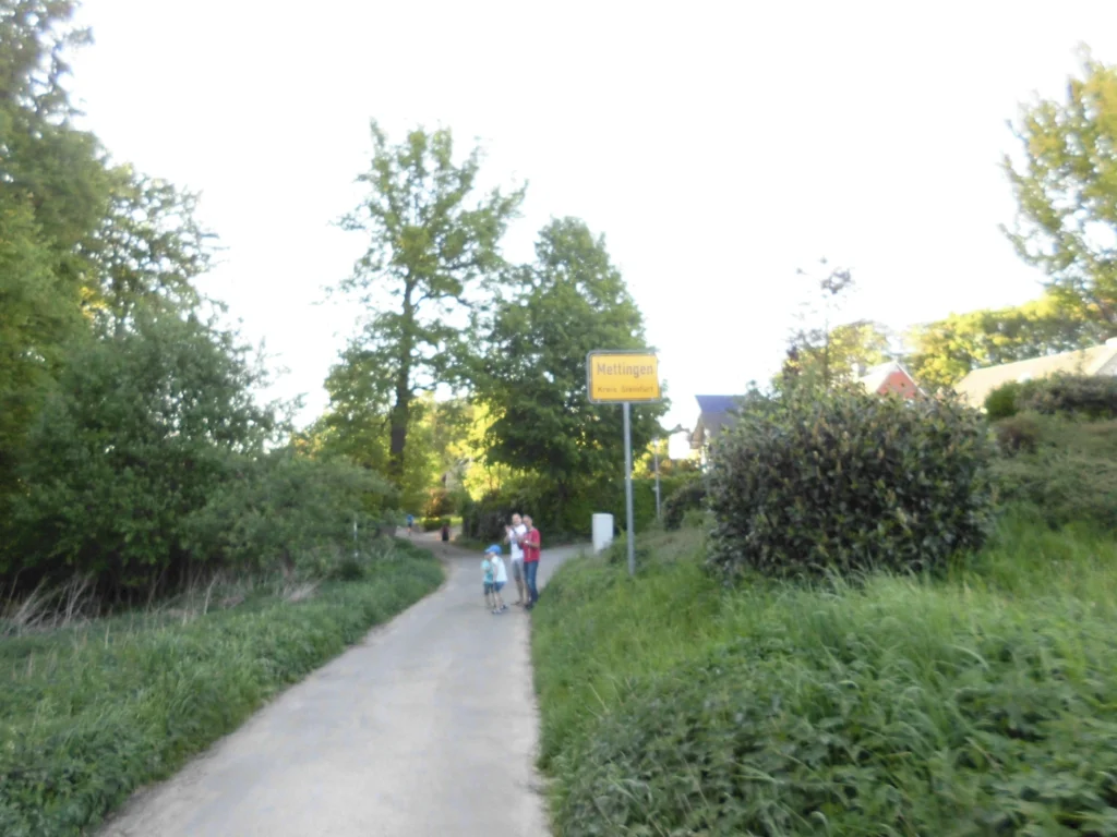 Weg in Mettingen, Nordrhein-Westfalen, umgeben von Bäumen und Sträuchern, mit einer Familie, die am Straßenrand steht. Der Ortsschild zeigt "Mettingen, Kreis Steinfurt".
