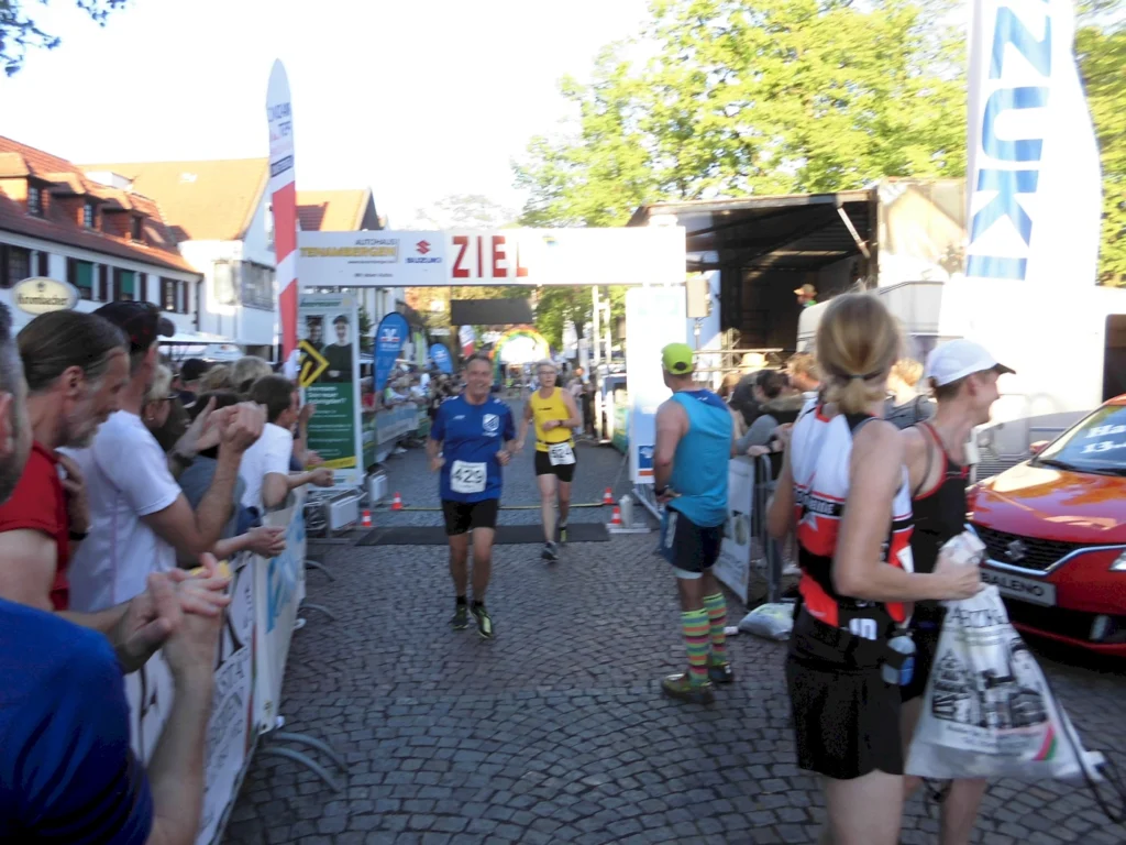 Läufer überqueren die Ziellinie bei einem Straßenlauf, umgeben von jubelnden Zuschauern. Im Hintergrund sind Werbebanner und ein Veranstaltungswagen zu sehen. Die Atmosphäre ist festlich und sportlich.