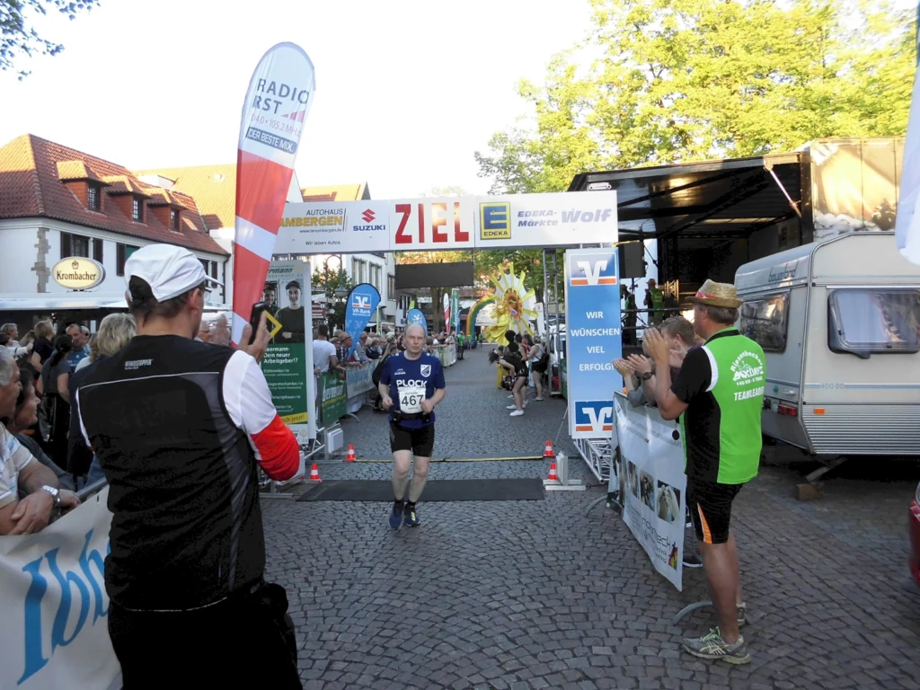 Ein Läufer mit der Startnummer 467 überquert die Ziellinie eines Laufwettbewerbs, während Zuschauer und Teilnehmer ihn anfeuern. Im Hintergrund sind Werbebanner und ein Wohnwagen zu sehen.