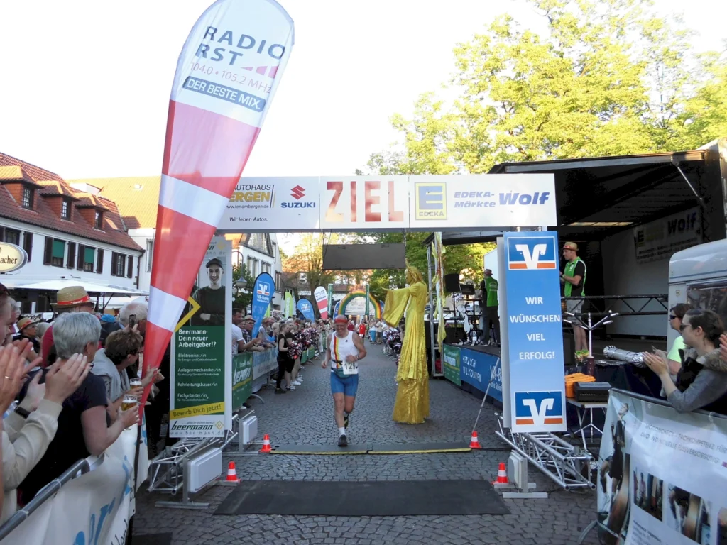 Läufer überquert die Ziellinie bei einem Straßenlauf, umgeben von jubelnden Zuschauern und Werbebannern. Im Hintergrund sind Sponsorenlogos sichtbar, darunter EDEKA und Suzuki. Die Veranstaltung findet in einer städtischen Umgebung bei sonnigem Wetter statt.