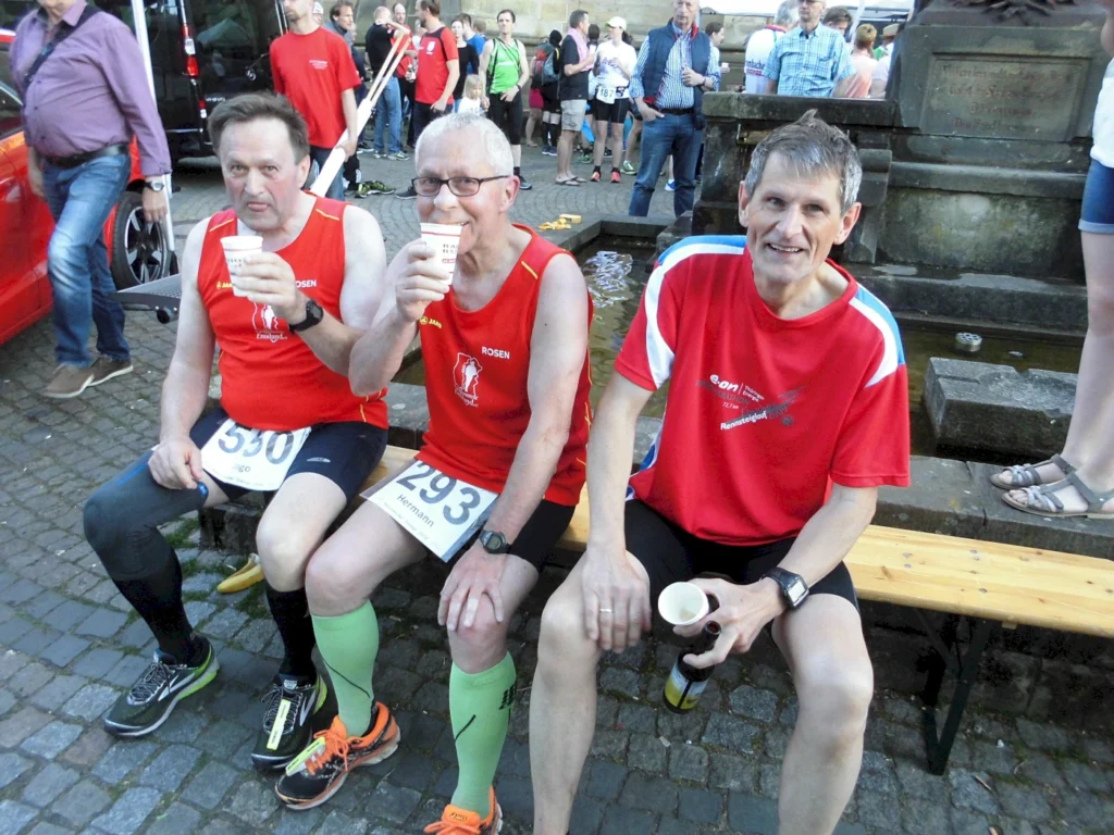 Drei Männer in roten Sporttrikots sitzen auf einer Bank, halten Becher in der Hand und genießen nach einem Lauf-Event eine Erfrischung. Die Umgebung zeigt eine belebte Veranstaltung mit Zuschauern im Hintergrund.