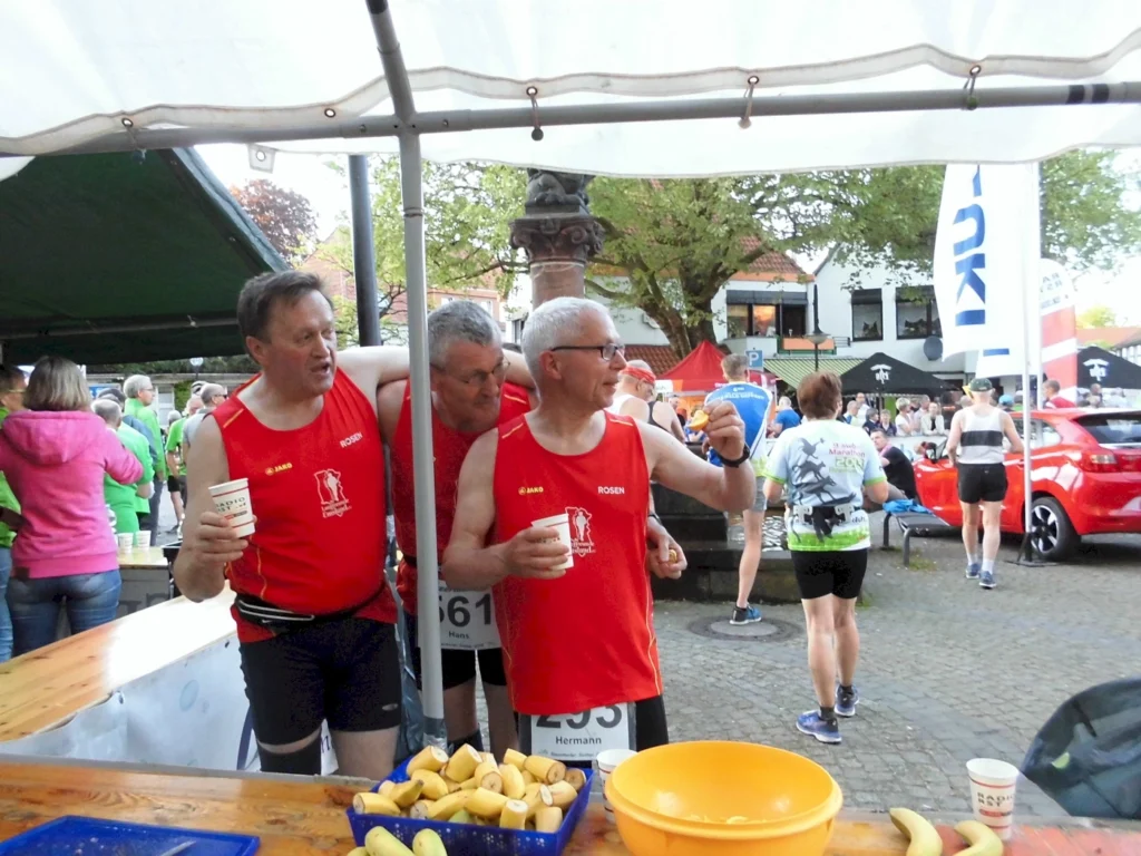 Drei Männer in roten Laufshirts stehen an einem Verpflegungstisch während eines Marathons, trinken aus Bechern und halten Bananen in der Hand. Im Hintergrund sind weitere Läufer und Zuschauer zu sehen.