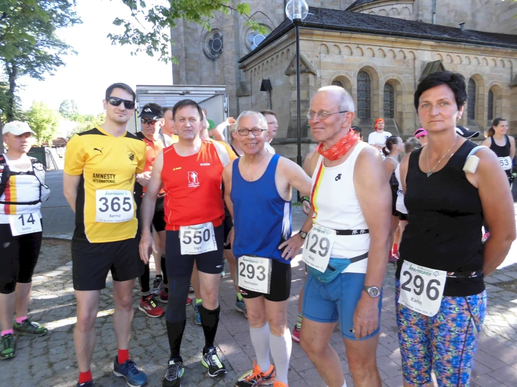 Läufer vor dem Start eines Wettlaufs, gekleidet in Sportkleidung und mit Startnummern. Die Gruppe besteht aus Männern und Frauen unterschiedlichen Alters, die sich auf das Rennen vorbereiten. Im Hintergrund sind Zuschauer und eine historische Kirche sichtbar.