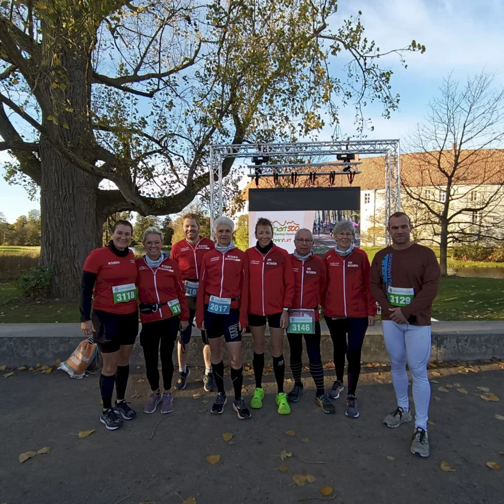 Gruppe von Läufern in roten Trikots vor einer Bühne mit Bildschirm, die an einem Lauf-Event teilnehmen, umgeben von herbstlichen Bäumen und einer malerischen Kulisse.