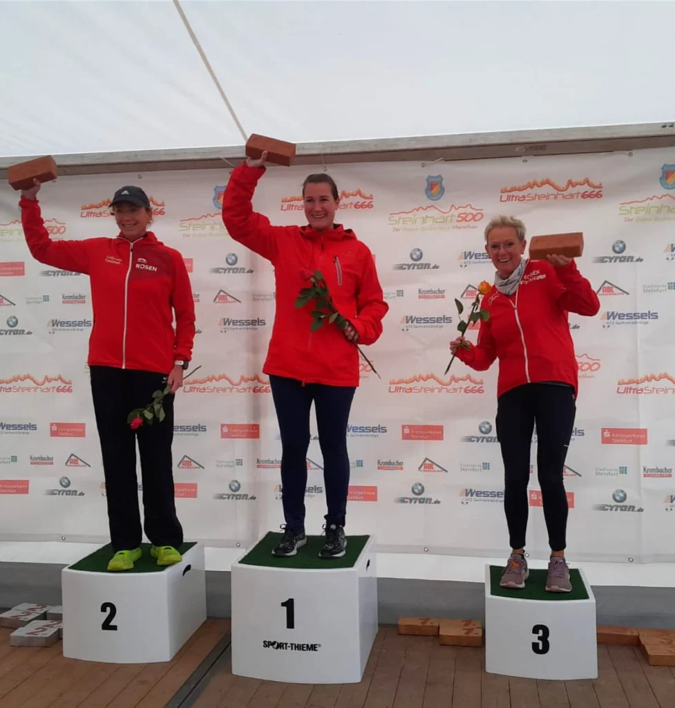 Drei Sportlerinnen stehen auf dem Siegertreppchen beim UltraSteinhart 666. Die erste Platzierte hält einen Pokal und eine Rose in der Hand, während die anderen beiden Sportlerinnen ebenfalls mit Blumen und Trophäen posieren. Alle tragen rote Jacken und zeigen Freude über ihre Erfolge.