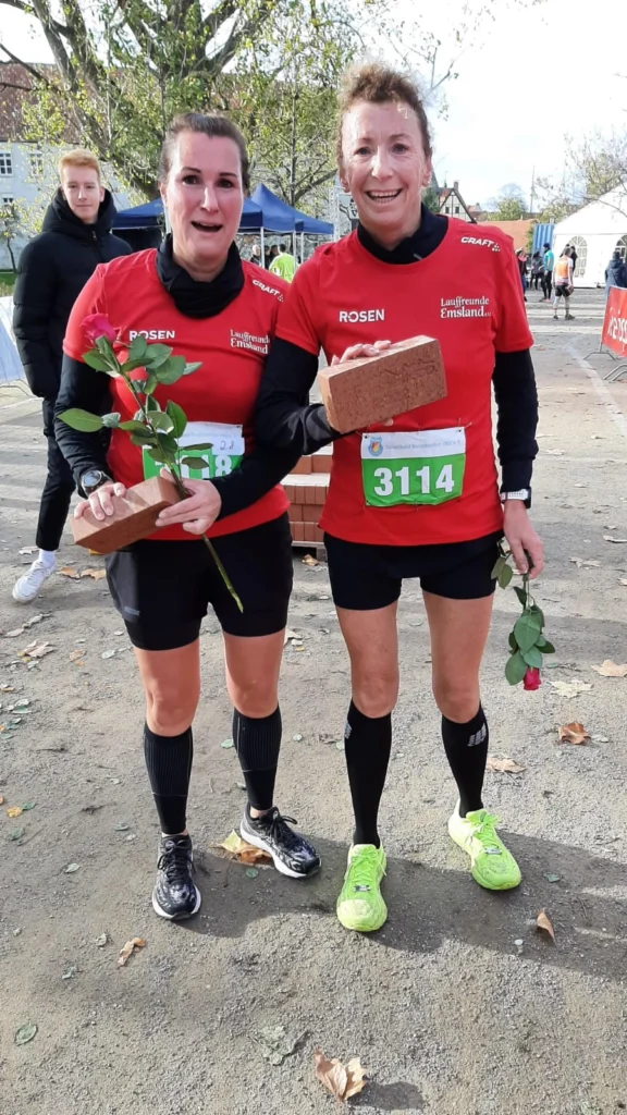 Zwei Frauen in roten Laufshirts der Lauffreunde Emsland halten stolz einen Ziegelstein und eine Rose nach dem Abschluss eines Laufevents. Im Hintergrund sind Zuschauer und Stände zu sehen.