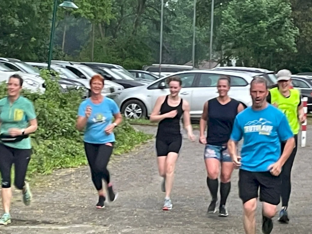 Gruppe von fünf Laufenden in Sportkleidung, die auf einem Weg joggen, umgeben von parkenden Autos und grüner Vegetation. Ideal für Themen wie Fitness, Laufen und gesundes Leben.