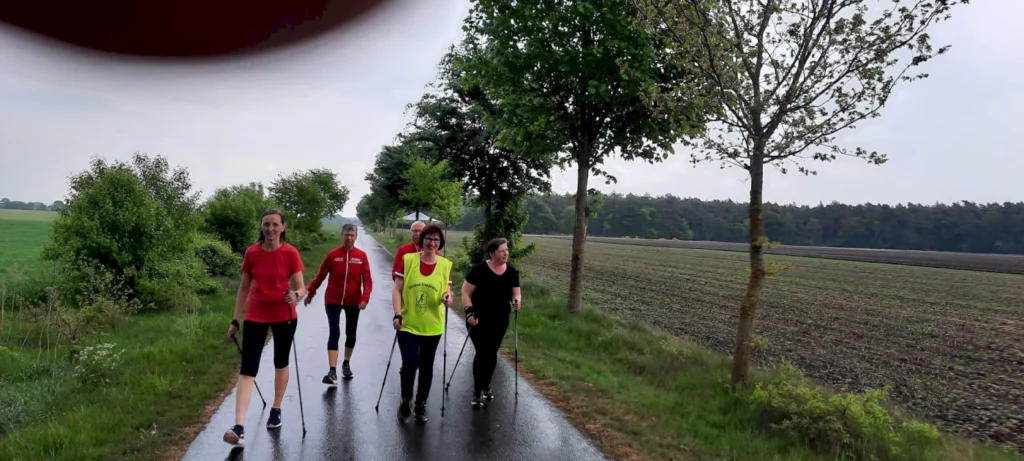 Gruppe von fünf Personen beim Nordic Walking auf einem ruhigen Weg, umgeben von Bäumen und Feldern, an einem bewölkten Tag.