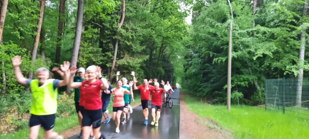 Läufer in bunten Sportoutfits joggen fröhlich auf einem Waldweg, umgeben von grünen Bäumen und frischer Natur. Einige Teilnehmer heben die Hände zur Begrüßung, während ein Radfahrer die Gruppe begleitet. Ideal für Laufveranstaltungen und Outdoor-Aktivitäten.