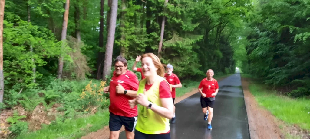 Gruppe von Läufern in roten T-Shirts, die auf einem Waldweg joggen. Die Umgebung ist grün und bewaldet, was eine aktive Freizeitgestaltung in der Natur zeigt.
