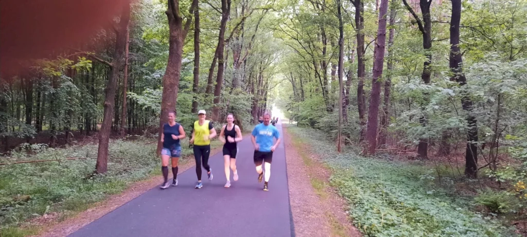 Gruppe von fünf Joggern auf einem Waldweg, umgeben von grünen Bäumen und Pflanzen, während sie in der Natur laufen. Ideal für Fitness und Outdoor-Aktivitäten.